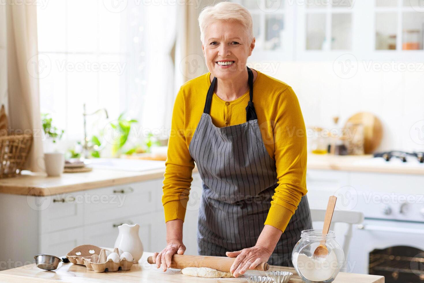 Attractive senior woman wearing apron making dough at kitchen, using rolling pin, copy space. Joyful elderly lady baking pastry cookies or pie at home. Domestic lifestyle, hobby in senior age concept photo