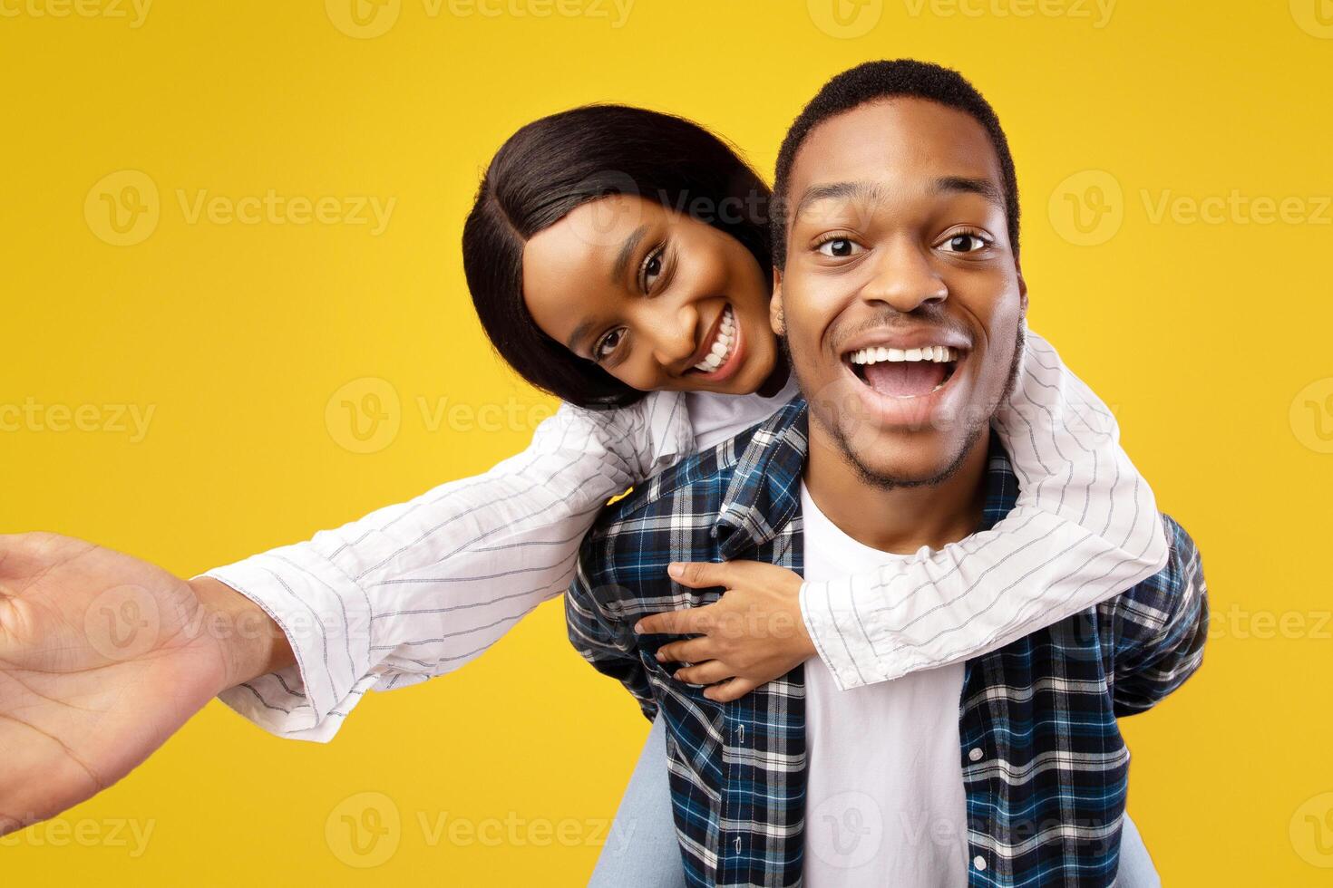 Portrait of lovers on date and have fun together. Happy young african american male holds woman on back, laughs with open mouth, lady makes selfie, isolated on yellow background, studio shot photo