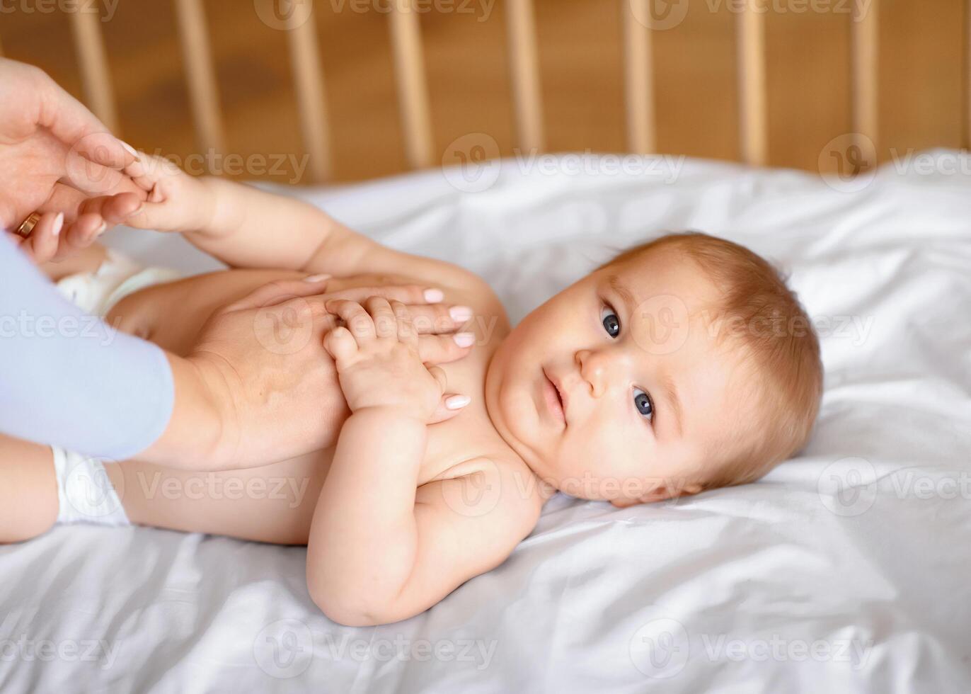 Unrecognizable mother massaging her cute little baby body. Closeup of adorable kid toddler looking at camera while playing with mom hands after waking up, laying in wooden baby bed photo