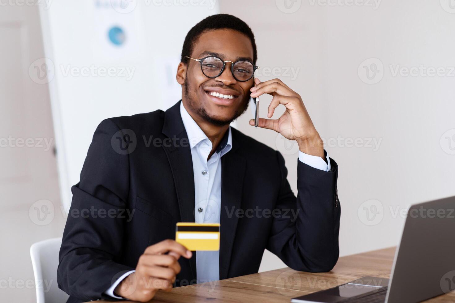 Easy payments. Happy african american businessman holding credit card after online purchase and talking on mobile phone with customer service while working with laptop in his office, free space photo