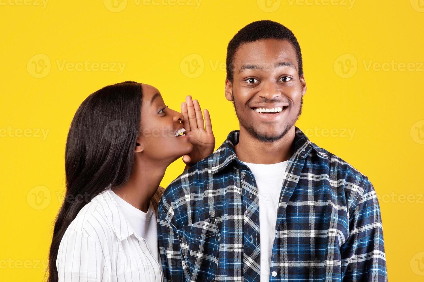 Big secret, sharing news. Mysterious millennial african american student lady in casual whispers in ear of smiling guy something interesting, isolated on yellow background, empty space, studio shot photo