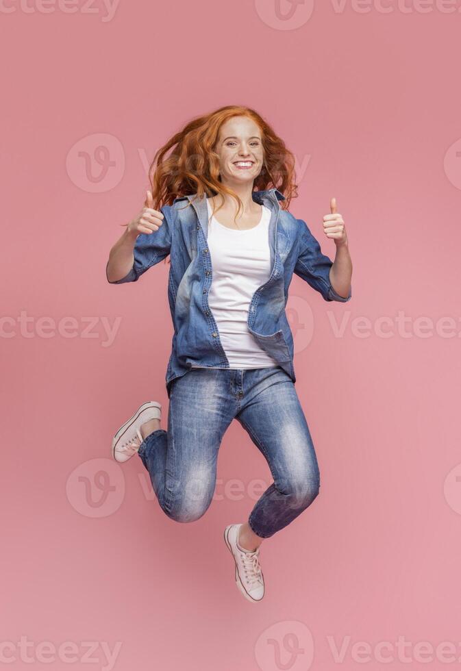 Happy young redhead teenager jumping and showing thumbs up, enjoy being in air. Pink studio background with free space photo