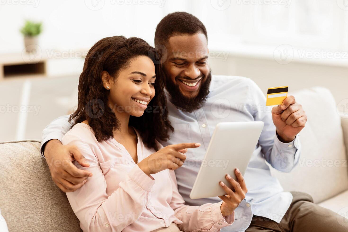 Easy Payment. Smiling positive young black couple holding debit credit card and using digital tablet, choosing and buying gifts, clothes or products, paying online, resting on couch in living room photo