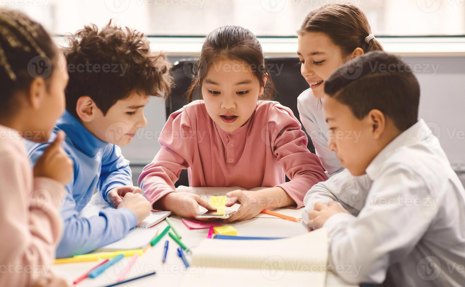 Diverse Group of Cute Small Children Using Smartphone, Sitting Together At Desk In Classroom Having Fun. Multicultural Kids Browsing Internet and Playing Online Games on Mobile Phone photo
