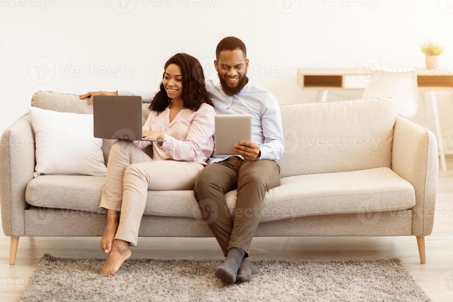 Weekend Leisure. Portrait Of Happy Millennial Black Couple Resting On Sofa. Man Using Digital Tablet, Woman Working On Laptop Computer. Family Browsing Internet Together Sitting On Couch photo