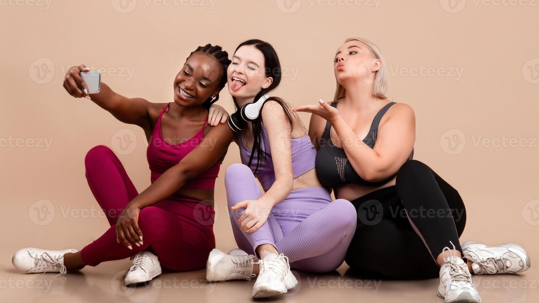 Cheerful Diverse Multiethnic Females In Sportswear Taking Selfie On Smartphone After Training, Happy Body Positive Females Having Fun Together, Sitting On Floor On Beige Studio Background, Free Space photo