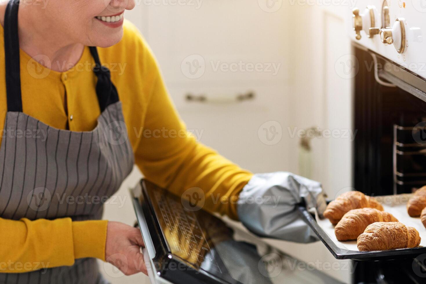 Closeup of smiling elderly woman baking pastry at home, taking delicious croissants out of oven, copy space. Joyful senior lady in apron opening oven, checking her pastry, cozy kitchen interior photo