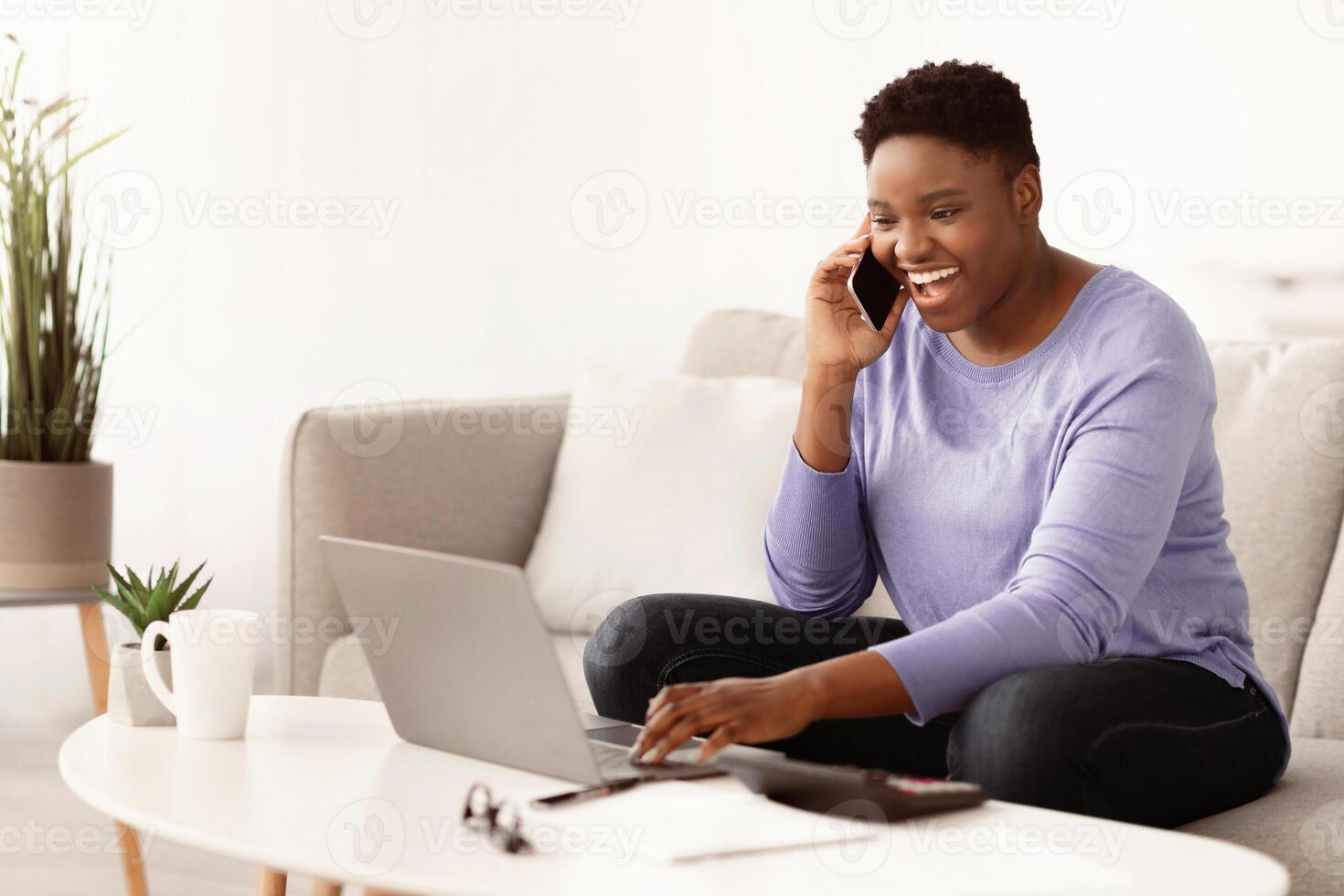 Informal Call. Emotional young curvy african american woman talking on her smartphone, sitting on the couch, using laptop, typing on computer keyboard. Copy space. Communication Concept. photo