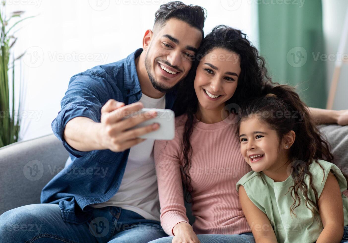 Family Selfie. Arab Man Taking Photo On Smartphone With His Wife And Little Daughter At Home, Happy Middle Eastern Family Posing At Camera Together While Sitting On Sofa In Living Room, Closeup
