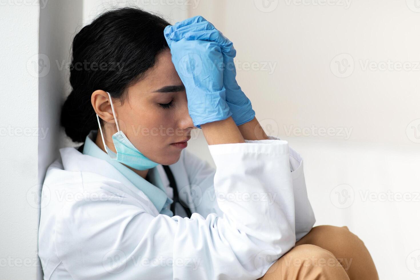 Tired female doctor sitting next to wall at clinic, wearing protective gloves and face mask, touching her forehead with hands, having difficult day, woman working hard during pandemic, copy space photo