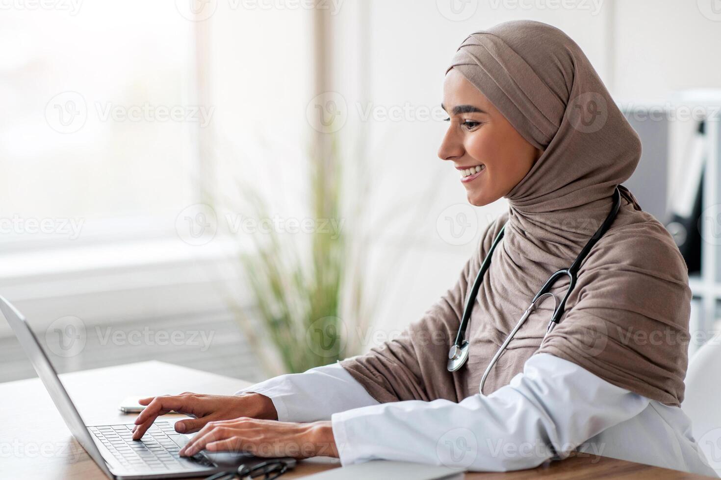 Young woman doctor having online training, sitting in her cabinet, using laptop and taking notes, side view, copy space. Attractive muslim lady physician working with computer, clinic interior photo