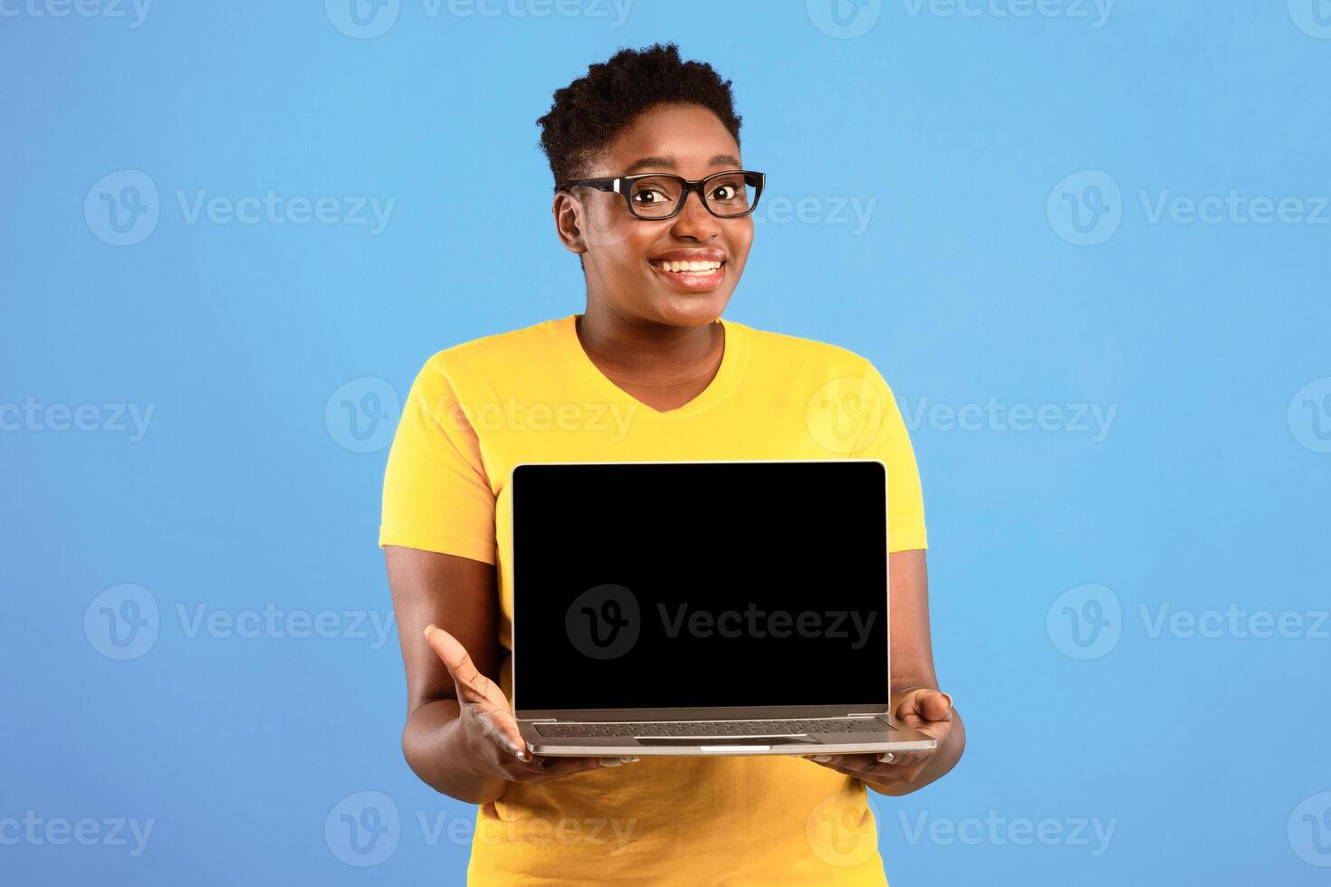 Smiling Oversized Black Woman Holding Laptop With Blank Screen Posing Over Blue Studio Background. Lady Showing Computer Monitor Recommending Website. Mockup photo