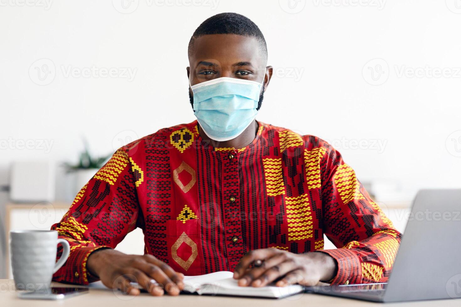 Portrait Of African Man In Medical Mask And Traditional Clothes Sitting At Desk In Home Office, Black Freelancer Guy Working Remotely During Coronavirus Pandemic, Looking At Camera, Copy Space photo