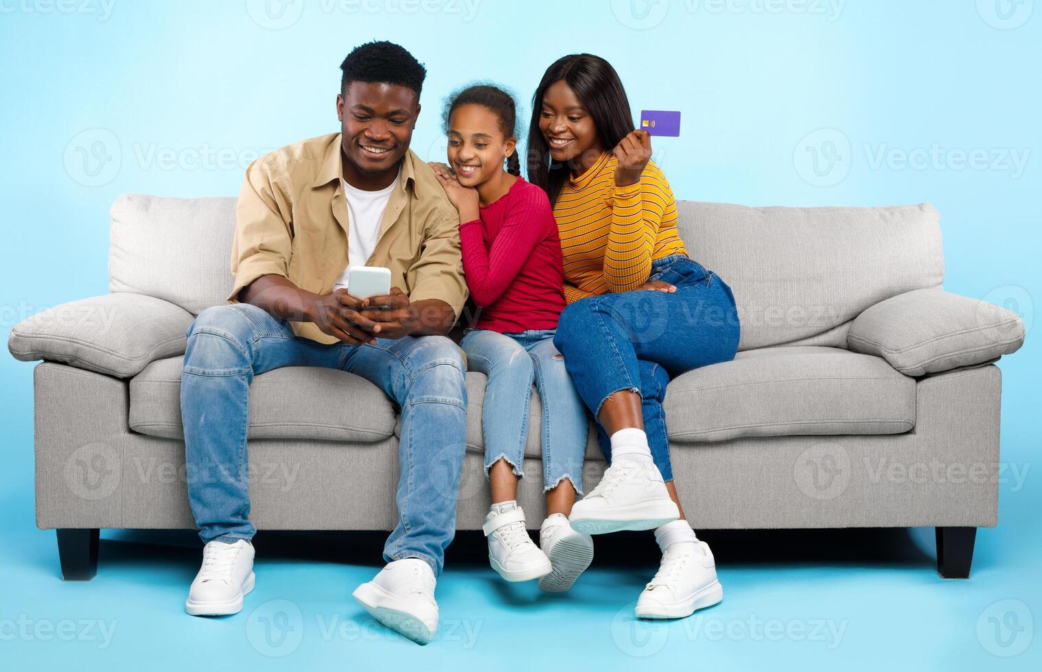 Online Shopping Concept. Happy Young Black Parents With Daughter Using Cell Phone And Debit Credit Card Buying Goods In Internet Web Store, Three People Sitting On Couch Isolated On Blue Studio Wall photo
