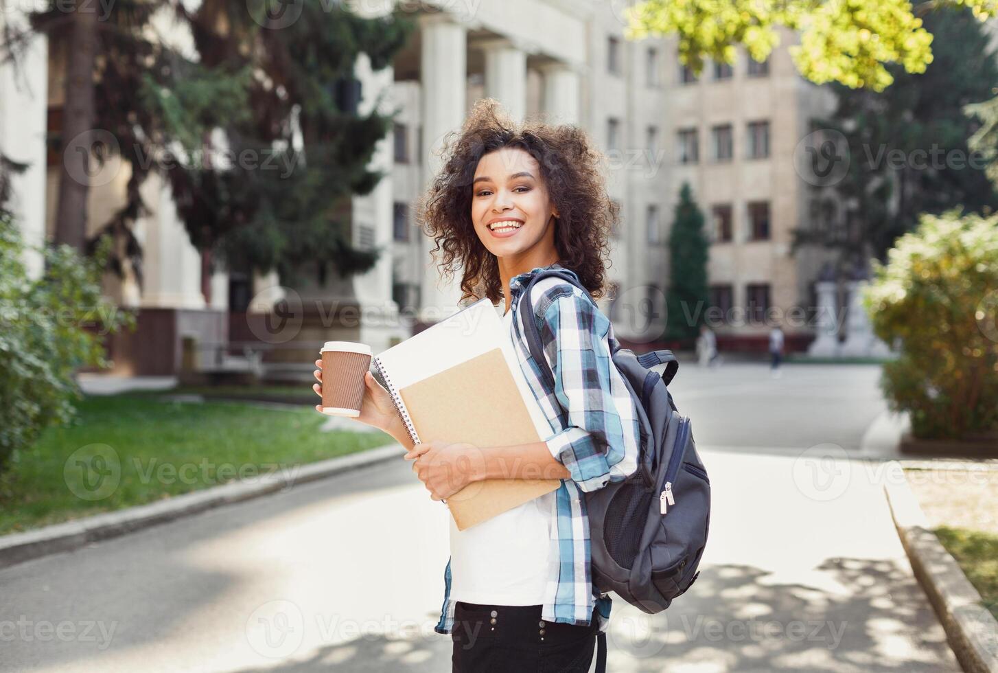 Happy smiling african-american student girl with backpack at university background. Woman with workbooks and take away coffee having rest in campus during lunch break. Education and leisure concept photo