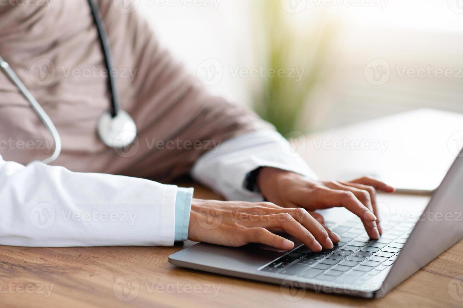 Cropped of female doctor having online training, sitting in her cabinet, using laptop, typing on keyboard, copy space. Unrecognizable muslim woman physician working with computer, clinic interior photo