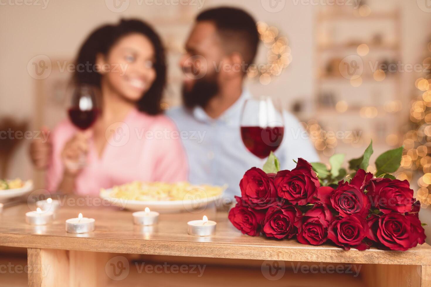 Romance And Bonding. Selective focus on bunch of red roses, bouquet of flowers lying on table. African american couple spending time together, having dinner, drinking wine in blurred background photo