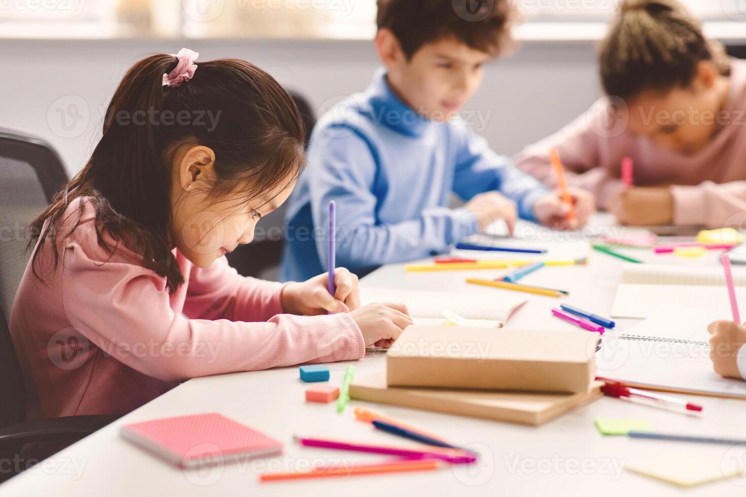 Portrait of small asian girl sitting at table in classroom at primary school or kindergarten, drawing in notebook with colorful pencils. Reopening and return back to school after lockdown photo