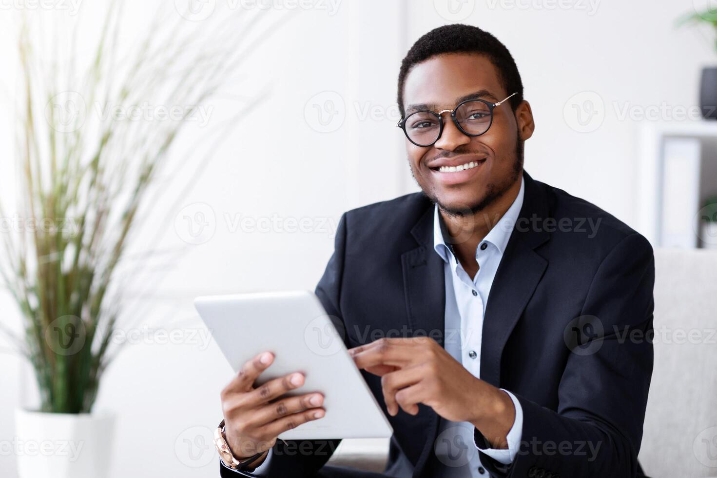 Smiling black manager in suit young man holding digital tablet, using modern technologies in business, office interior, copy space. Cheerful african american businessman using app for business photo