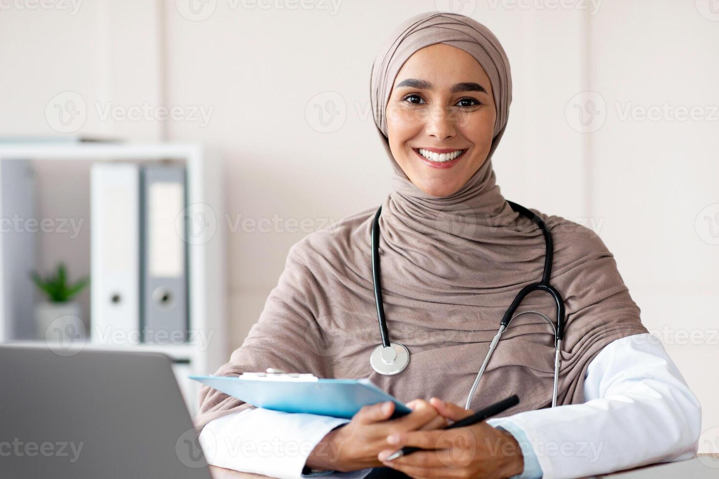 Smiling young woman doctor in hijab taking anamnesis in hospital, sitting in front of laptop, holding medical chart and pen, copy space. Female therapist in head scarf smiling at camera while working photo