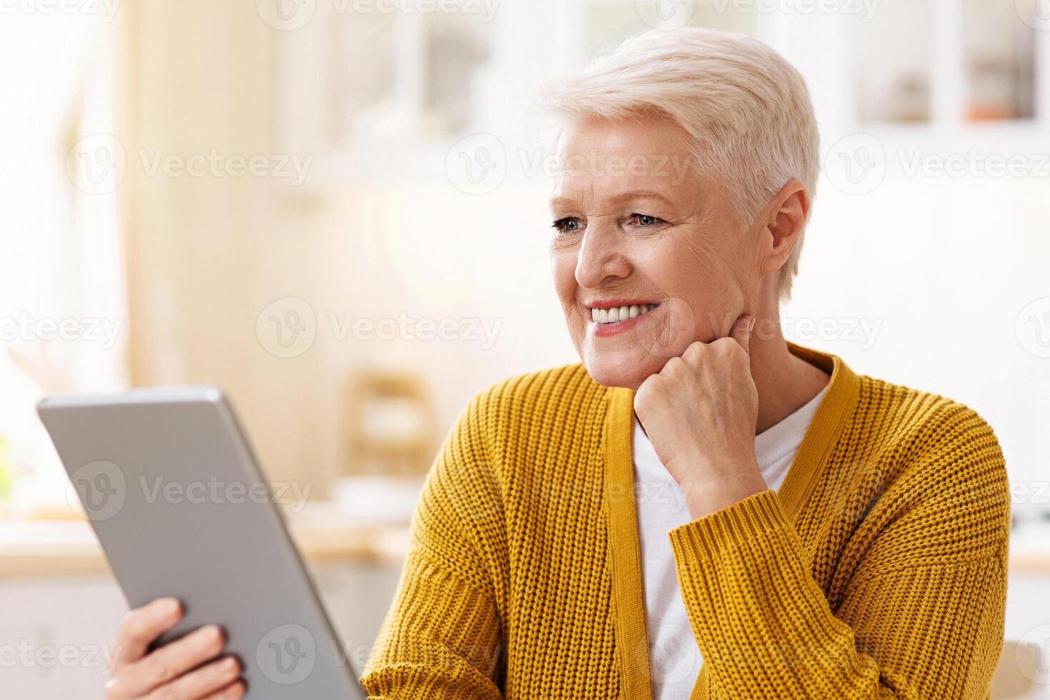 Joyful old grey-haired woman drinking coffee and using digital tablet in kitchen, empty space. Happy grandmother reading culinary blog or book online, surfing on Internet on modern pad at home photo