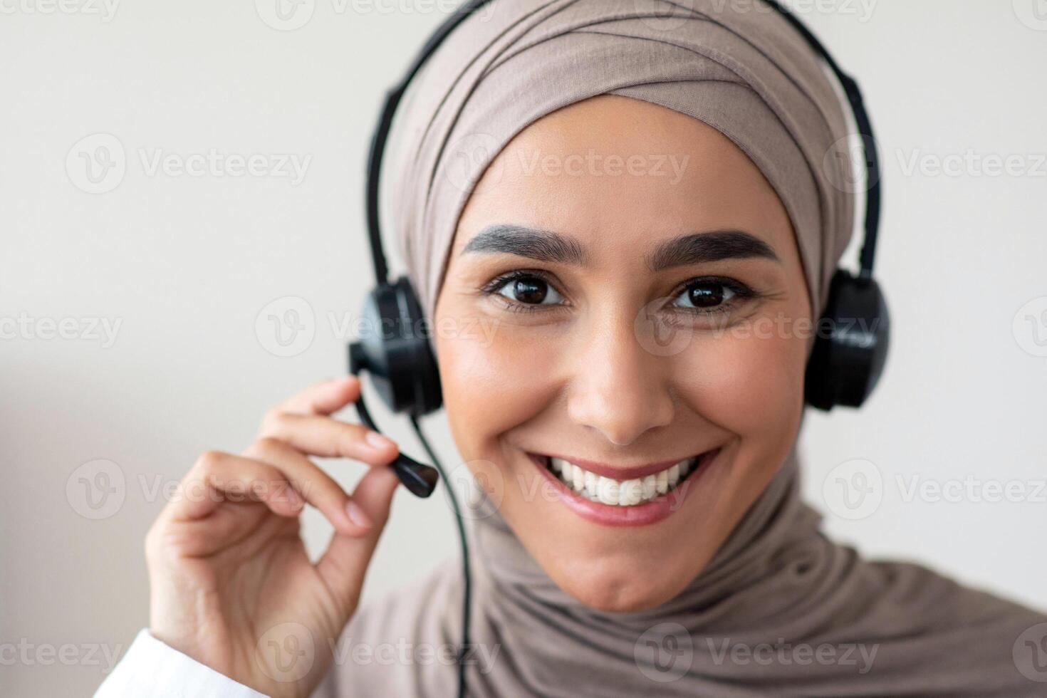 Closeup portrait of cheerful female doctor muslim woman with headset talking to patient, copy space. Smiling young lady in hijab having conversation with patient, touching microphone photo