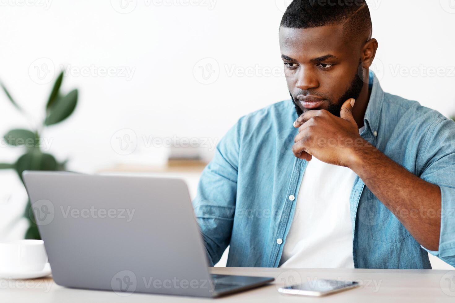 Focused African American Businessman Working With Laptop At Office, Sitting At Desk And Looking At Computer Screen, Black Entrepreneur Thinking About Project Strategy And Business Plan, Copy Space photo