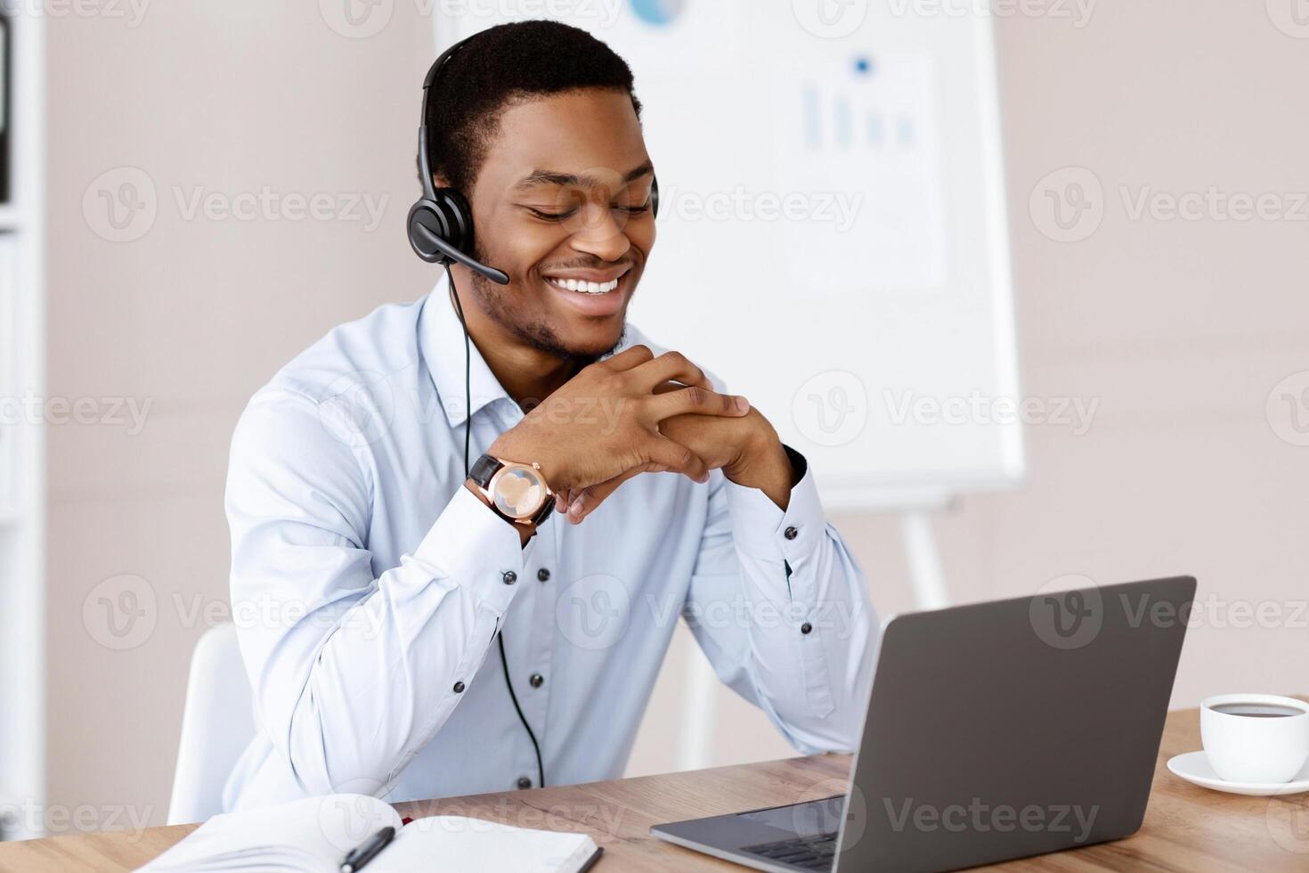 Happy black man having online training, using headset and laptop, looking at computer screen and smiling, office interior, copy space. Cheerful african guy having online call with clients photo