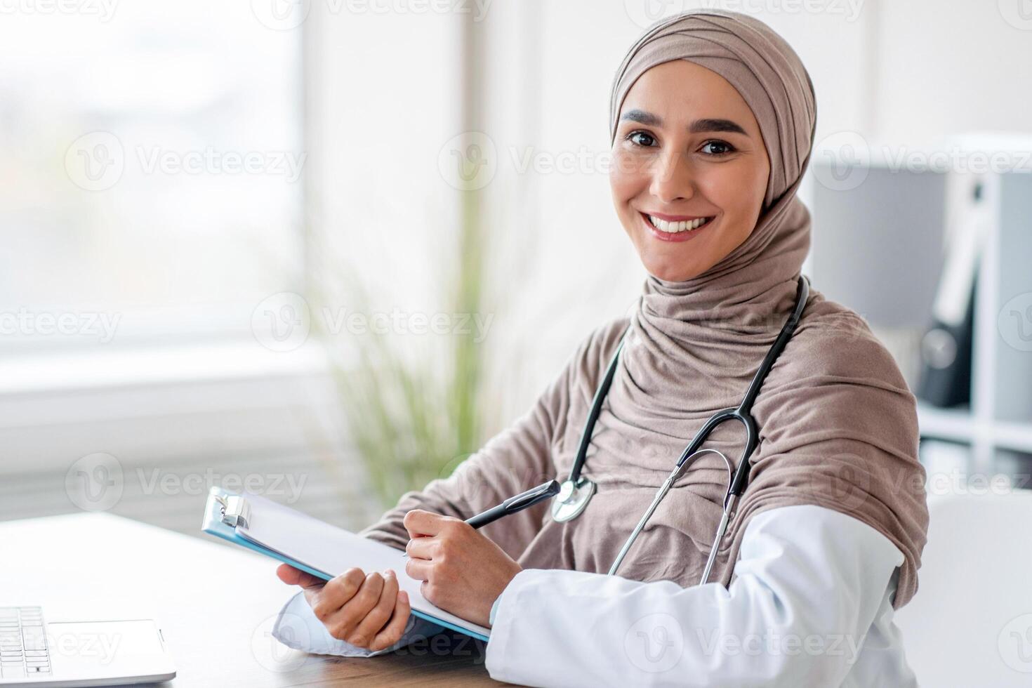 Female doctor taking anamnesis while having conversation with patient. Smiling muslim woman doctor in headscarf taking notes in medical chart, sitting at workdesk at her cabinet in clinic, copy space photo