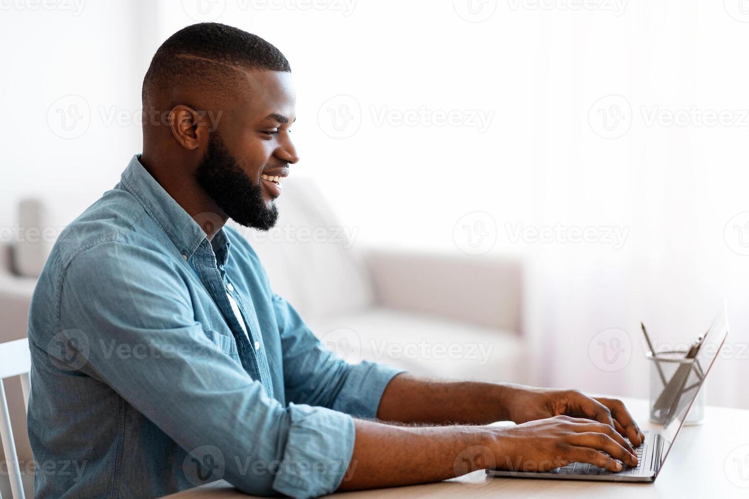 Smiling Millennial African American Man Working With Laptop At Home Office. Handsome Black Self-Employed Guy Typing On Laptop Keyboard, Positive Male Freelancer Sitting At Desk, Side View, Copy Space photo
