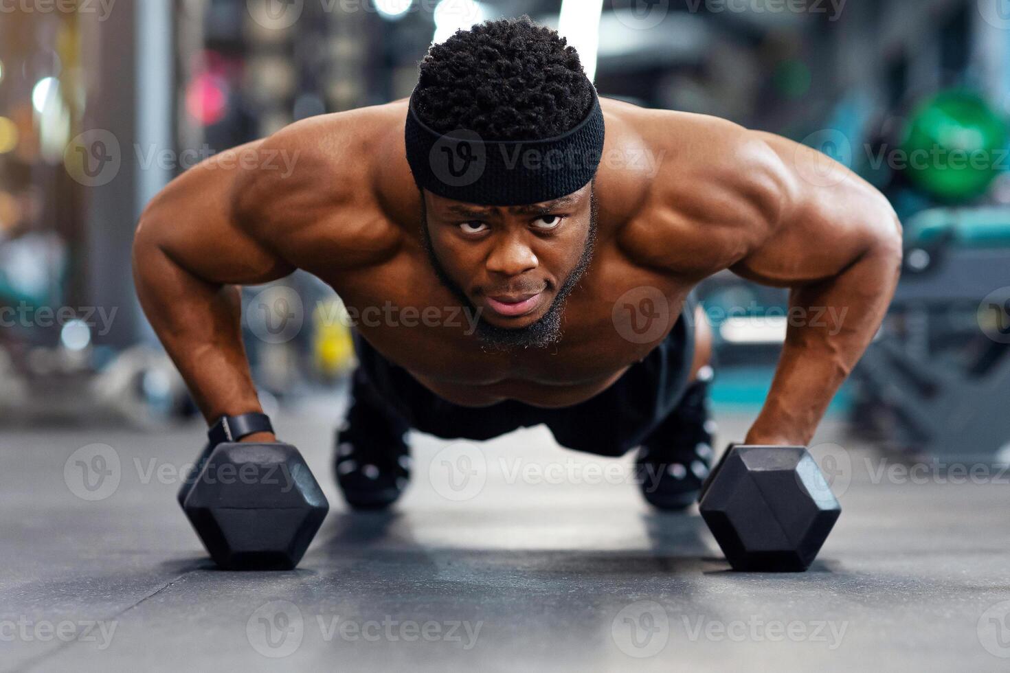Confident black guy sportsman pushing up with dumbbells, looking at camera, panorama. Muscular african american shirtless man holding barbells and pushing from floor, work out at gym photo