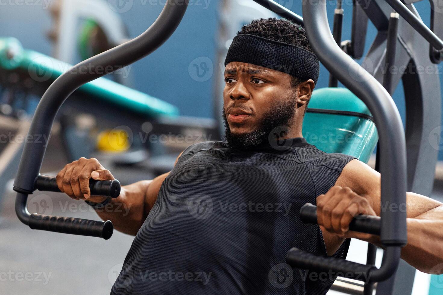 Motivated young african american man having workout on chest press machine in gym, pushing weight while sitting on bench, building up arms and torso muscles, closeup, looking at copy space photo