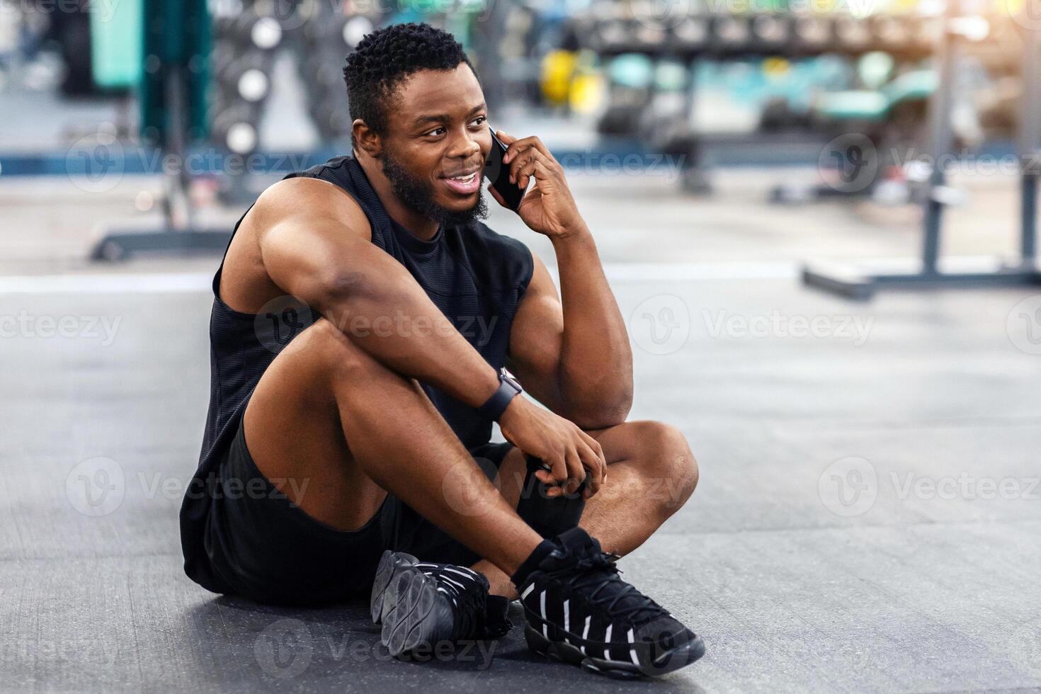 Smiling african american athletic man resting after workout in gym, copy space. Muscular black guy sitting on floor in gym, holding bottle with protein or water and talking on mobile phone photo