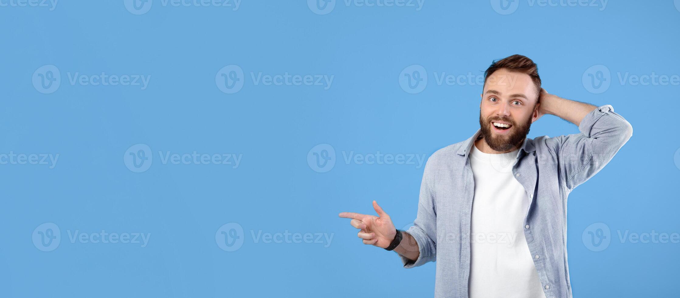 Happy young guy pointing at empty space, cannot believe his luck over huge sale on blue studio background, banner design with copy space. Handsome bearded man promoting your product photo