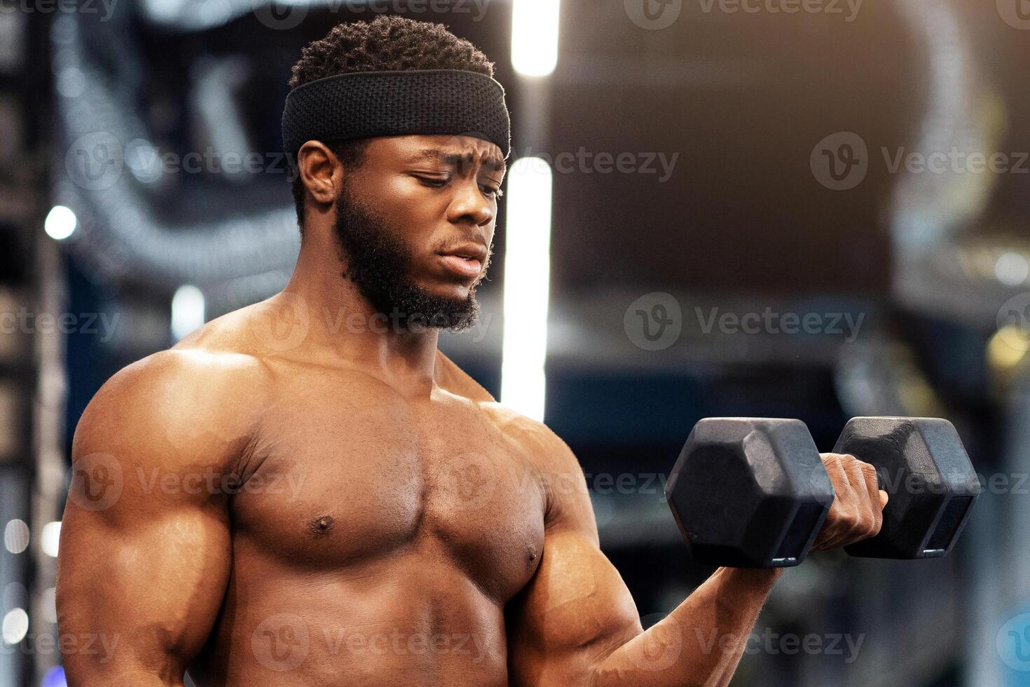 Concentrated handsome african american shirtless bodybuilder making work out with barbells at gym, looking at copy space. Hot black guy exercising with two dumbbells, working on his muscular biceps photo