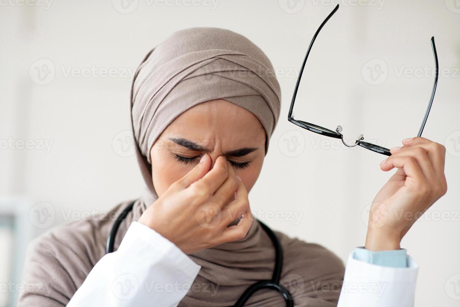 Closeup of female doctor woman in hijab removing glasses and rubbing nose bridge, suffering from tired eyes syndrome, clinic interior. Eye fatigue, symptoms and treatments concept photo