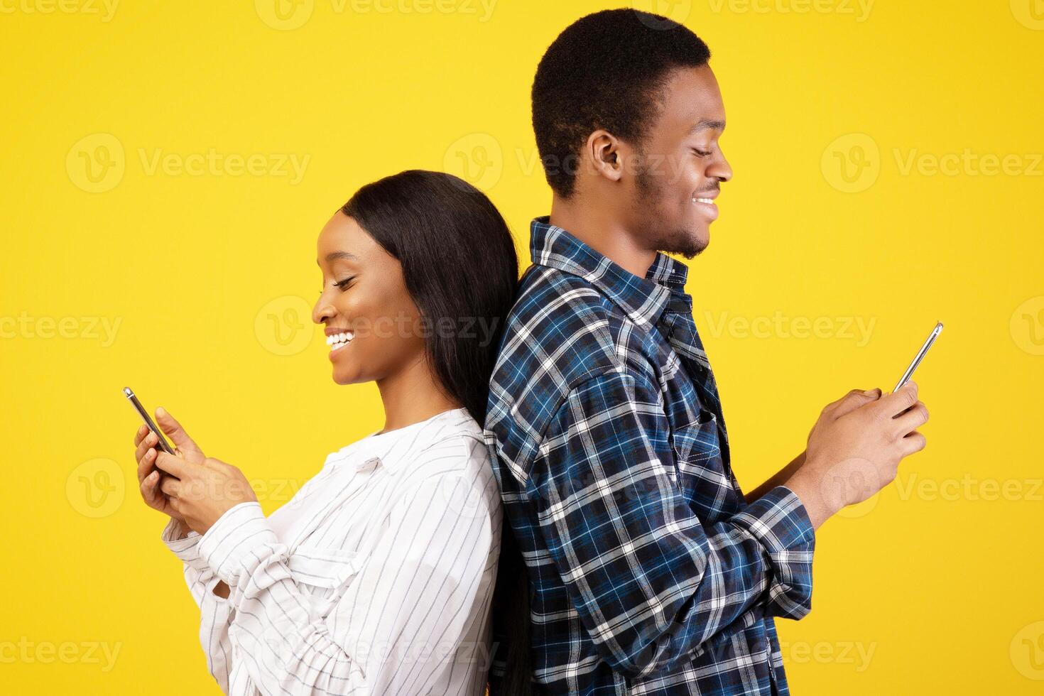 Social network and modern online relations. Smiling young african american guy and lady stand back to back, typing on smartphones, isolated on yellow background, studio shot, profile, copy space photo