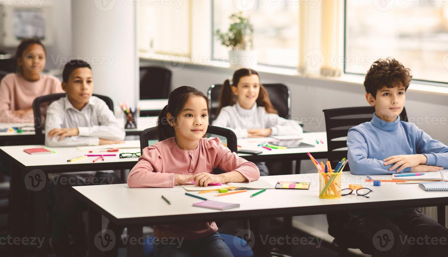 Back To School. Portrait of focused multicultiral pupils sitting with folded arms at desks with social distance, listening to teacher. Diverse junior students working diligently and learning new stuff photo
