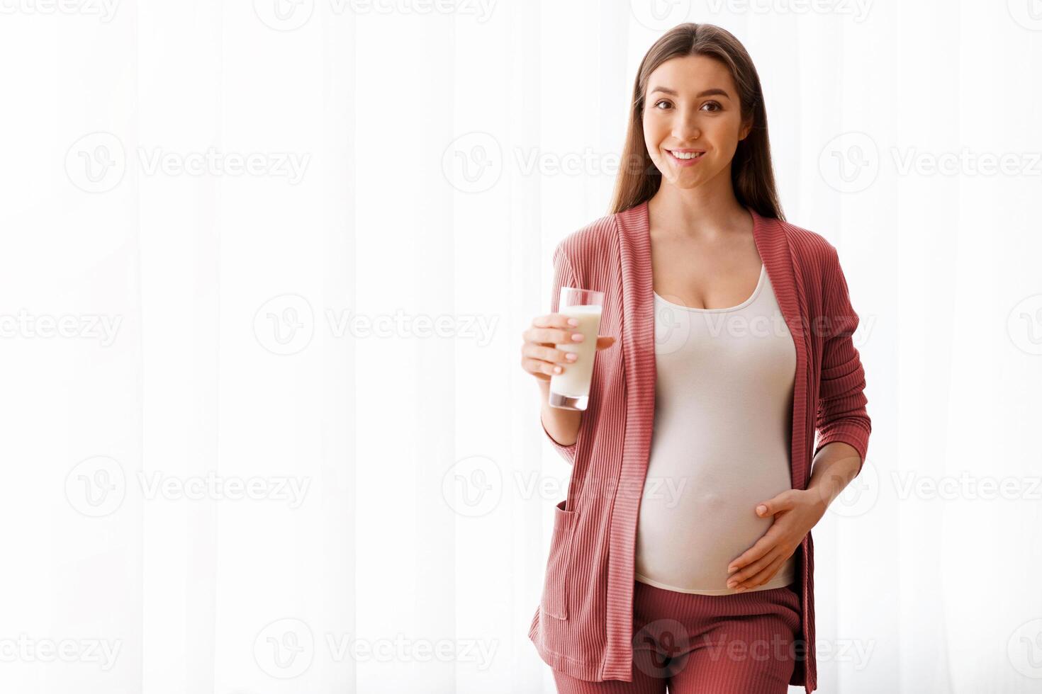 Beautiful Smiling Pregnant Lady With Glass Of Milk Posing Near Window At Home, Positive Millennial Expectant Woman Choosing Healthy Calcium Drink During Pregnancy And Looking At Camera, Copy Space photo