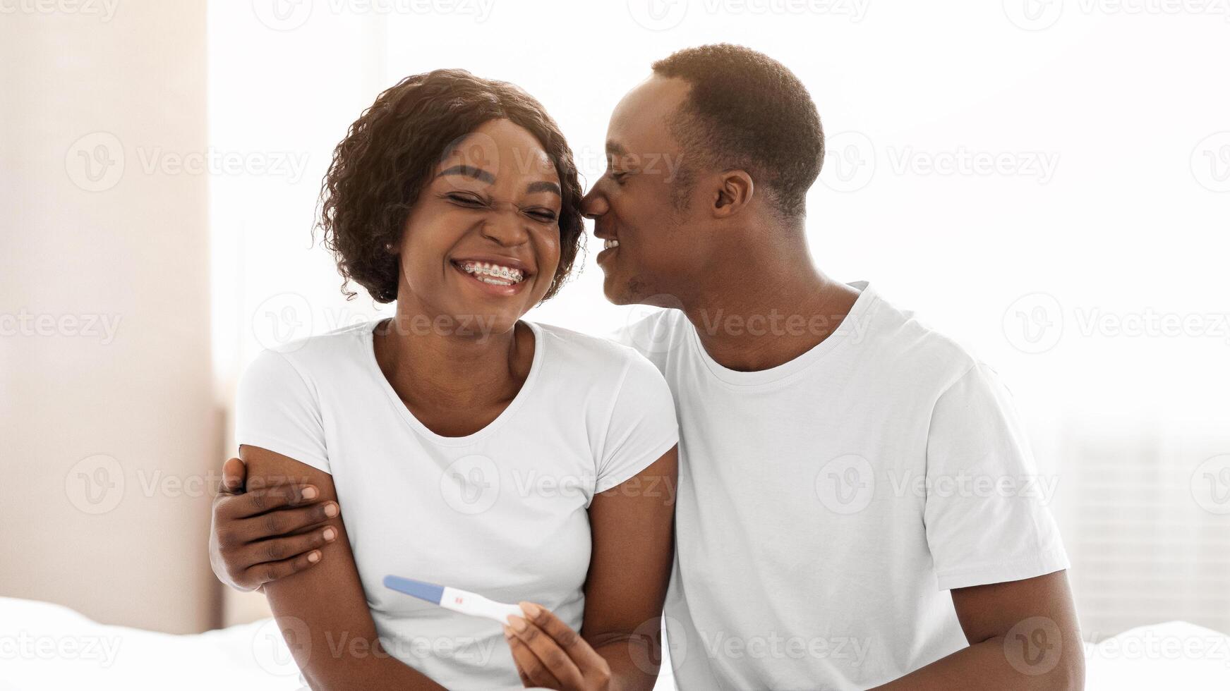Emotional black couple future parents holding positive pregnancy test, husband hugging his happy wife, bedroom interior, empty space. Delighted african american family waiting for baby photo