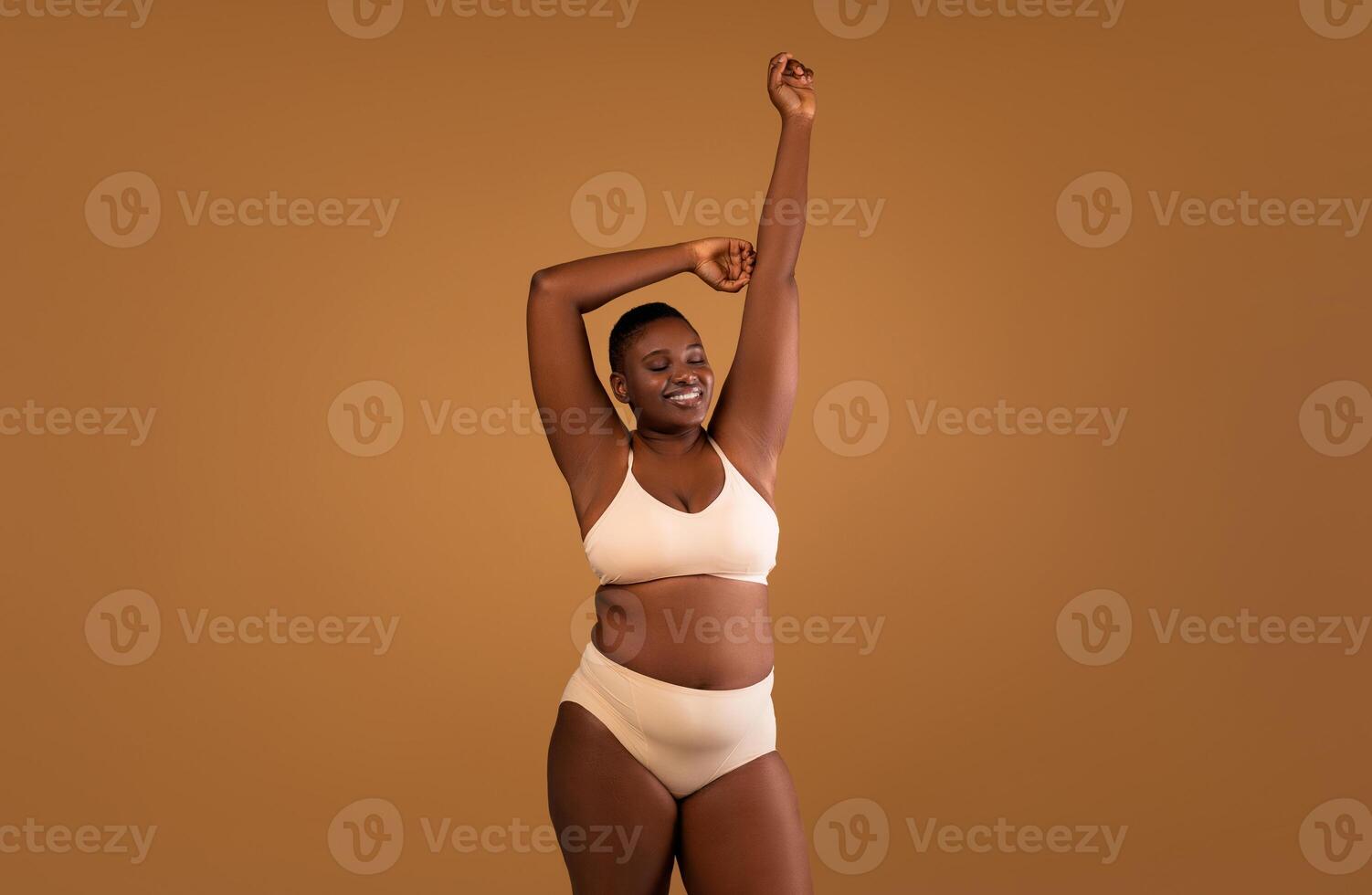 Portrait Of Beautiful Curvy African American Lady Posing Isolated On Brown Studio Background, Black Lady In Beige Underwear Enjoying Herself, Standing With Raised Arms And Closed Eyes, Free Copy Space photo