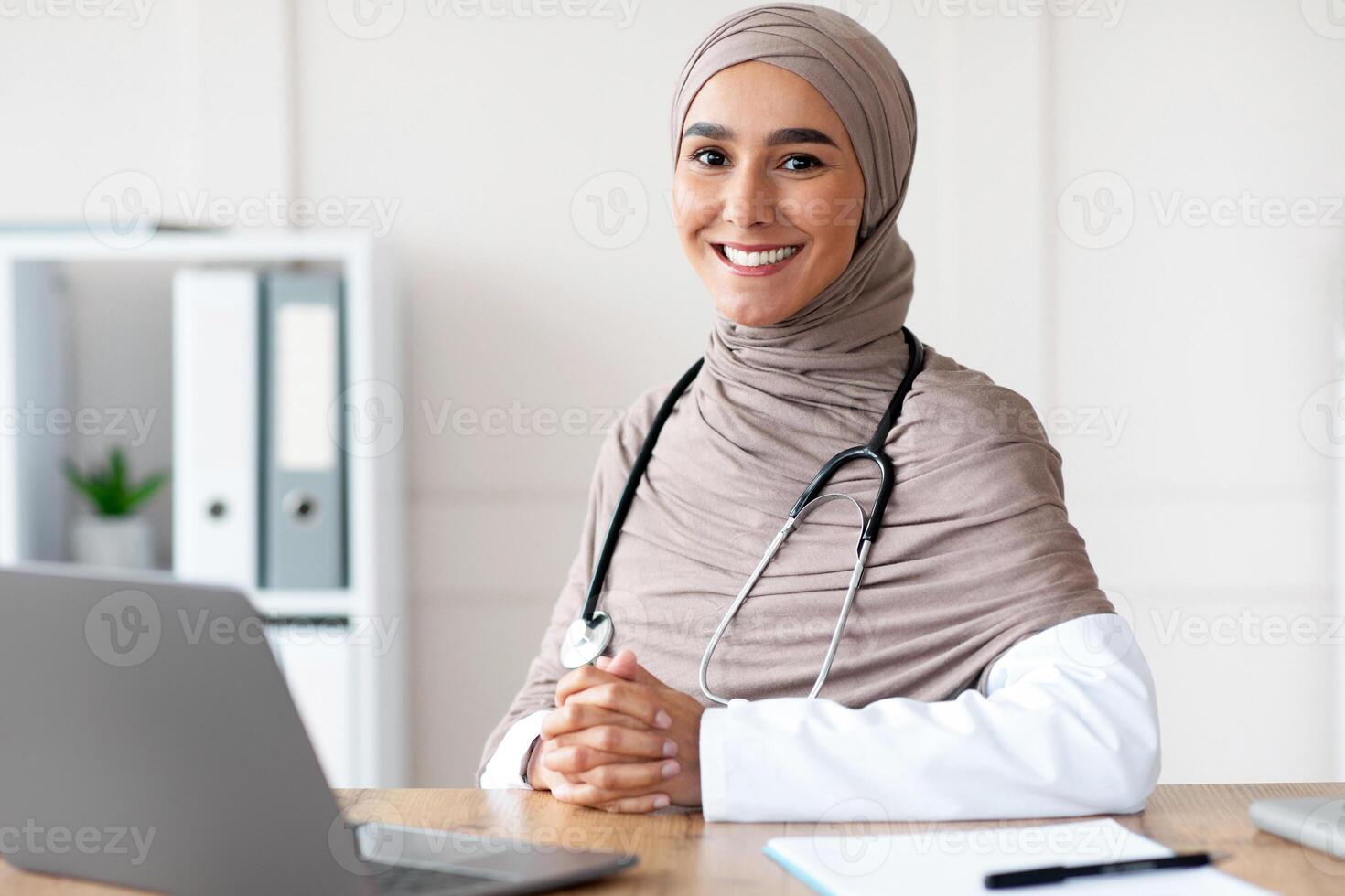 Smiling female doctor muslim woman posing in her cabinet at clinic, wearing hijab, sitting in front of laptop, having medical chart on her table, working online or taking patient anamnesis, free space photo