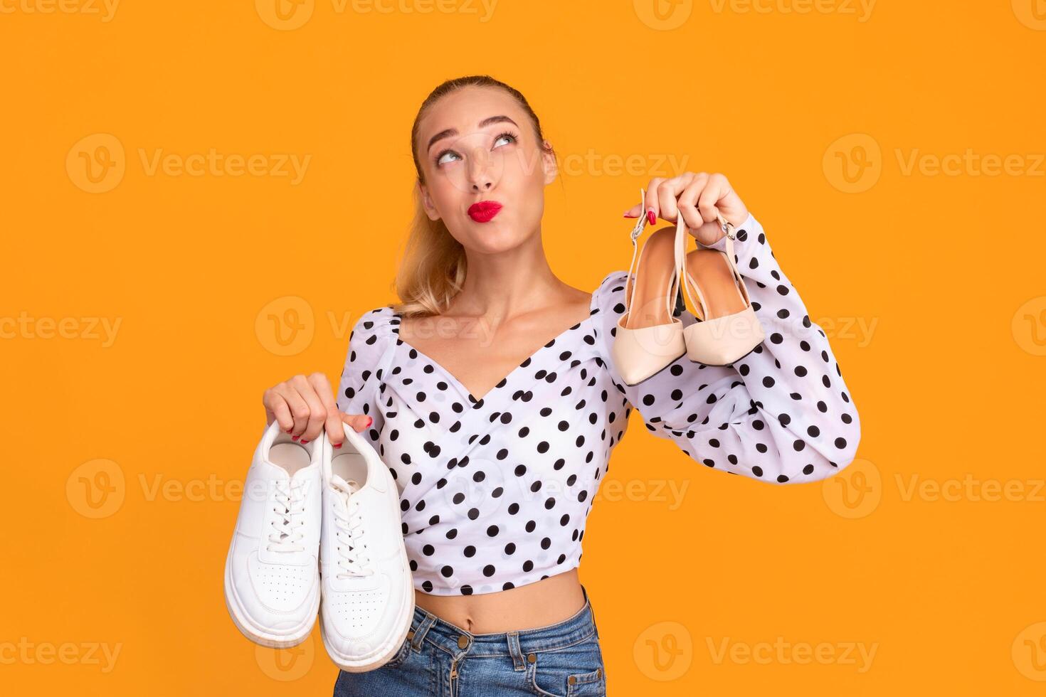 Elegance Or Sport. Funny pensive woman holding pair of high heels shoes and white sneakers in hands, thinking and choosing what to wear, looking up, isolated over yellow background wall, free space photo