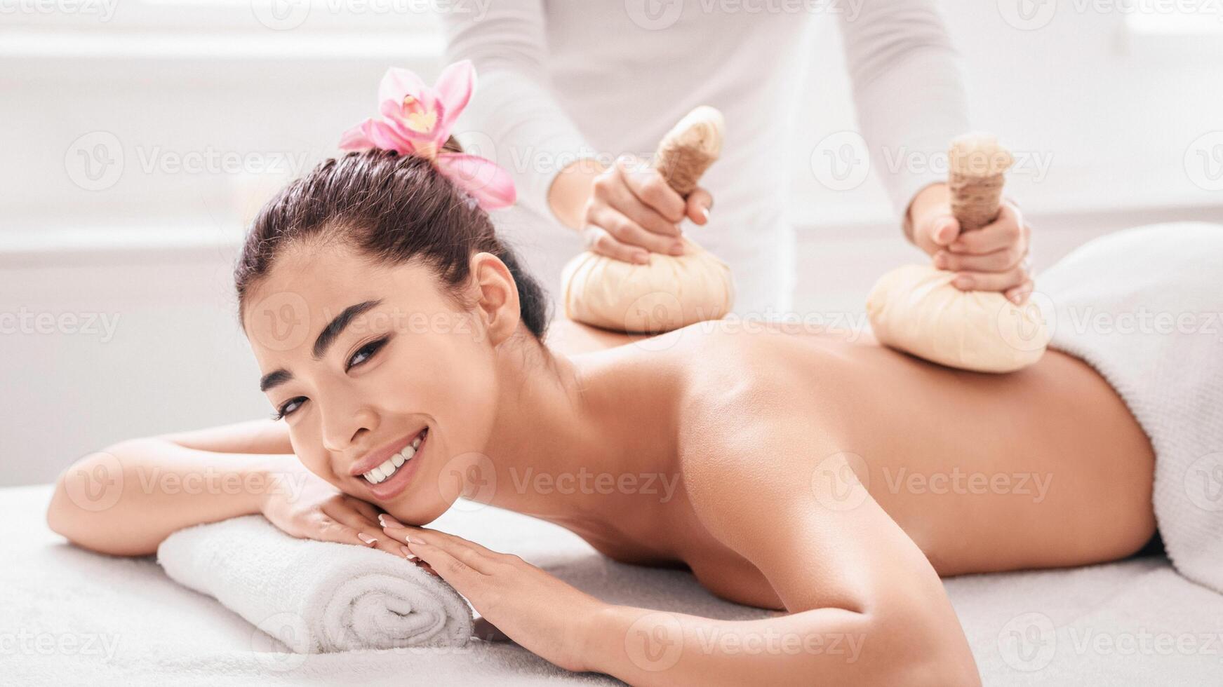 Pampering Day At Spa. Young Asian Woman Enjoying Massage With Herbal Balls, Attractive Korean Lady Having Wellness Beauty Treatment In Salon, Lying On Table, Looking And Smiling At Camera, Closeup photo