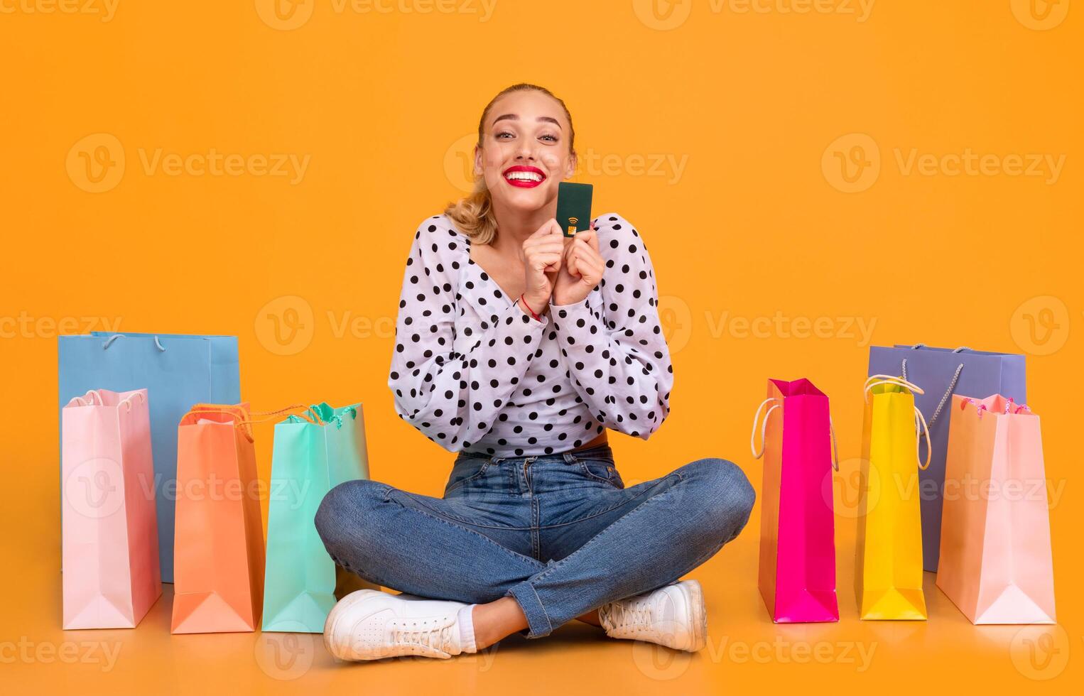 Easy Payment. Portrait of excited stylish young woman holding credit card, ordering purchases and paying online via internet, buying clothes, sitting at yellow studio on the floor with shopping bags photo