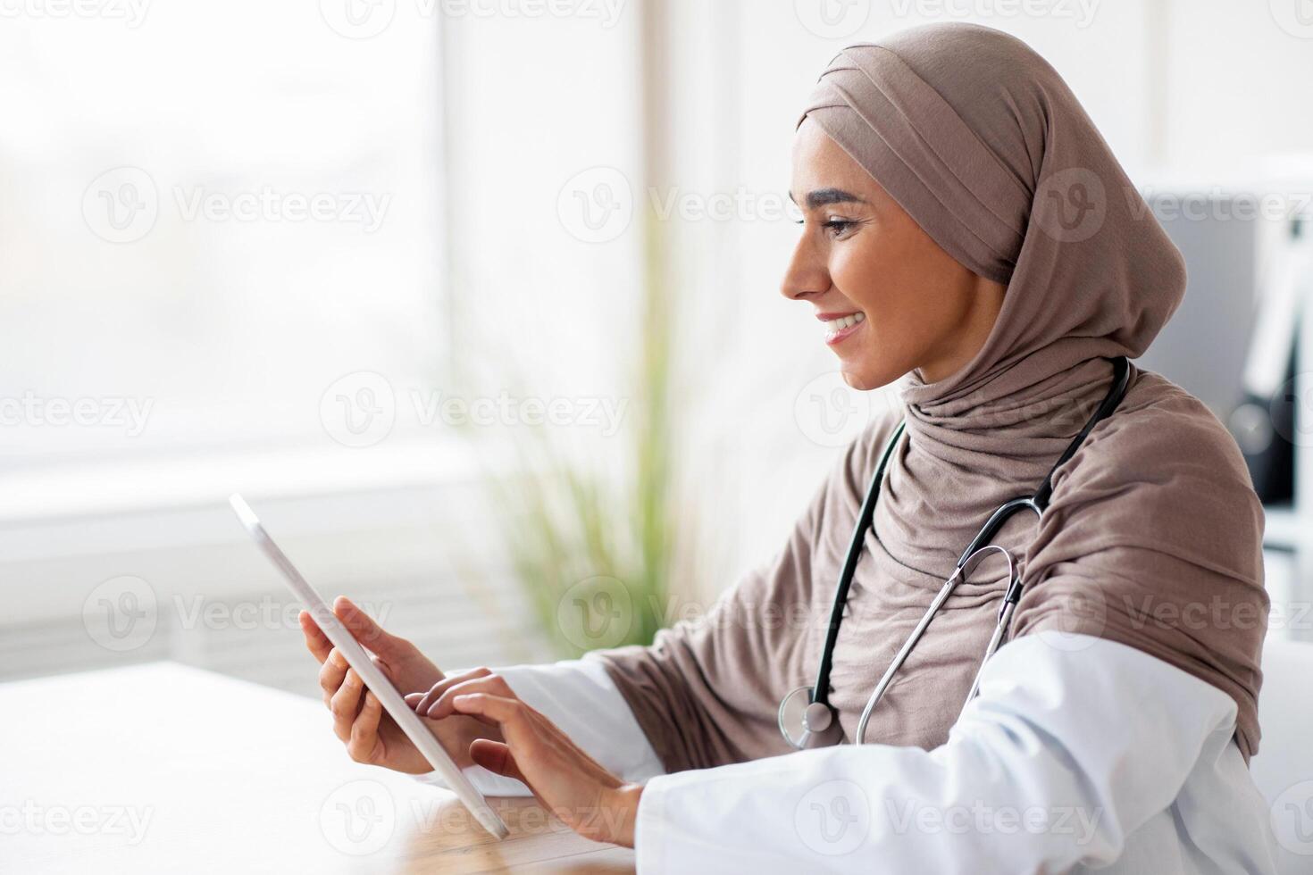 Female doctor muslim woman using digita tablet while working in clinic or hospital, having online appointment with patient, using application, reading medical articles, side view, copy space photo