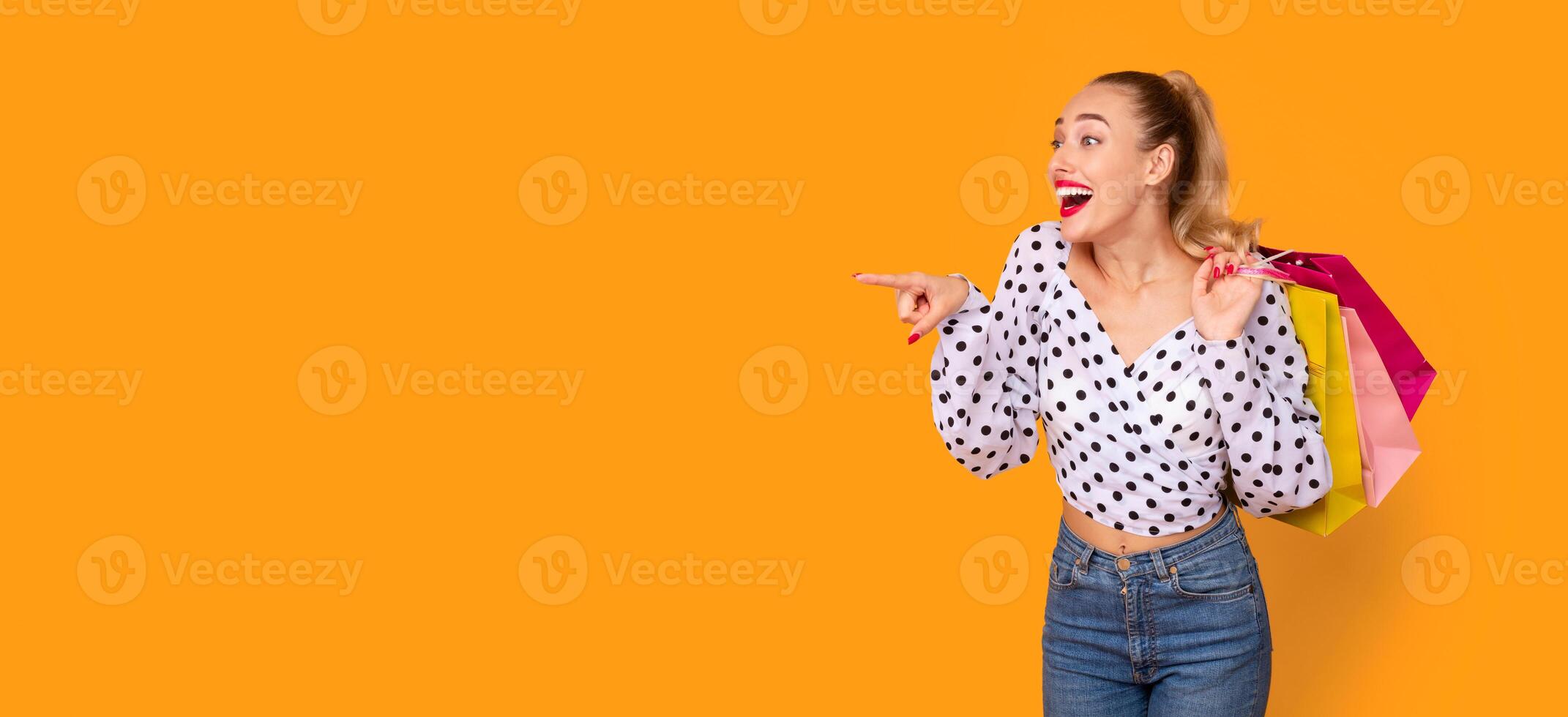 Wow, Look At It. Portrait Of Excited Stylish Woman Pointing Finger Aside At Free Space For Promotional Text Or Offer, Carrying Colorful Shopping Bags Isolated On Orange Studio Background, Panorama photo