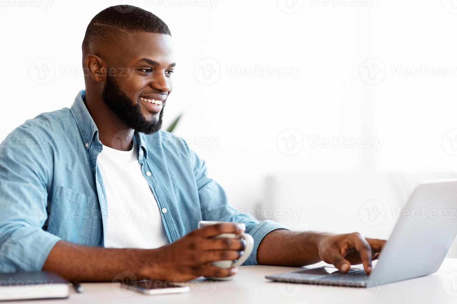 Freelancing Concept. Cheerful Black Freelancer Man Drinking Coffee And Working On Laptop At Home Office, Millennial African American Guy Sitting At Desk With Computer, Enjoying Remote Job, Copy Space photo