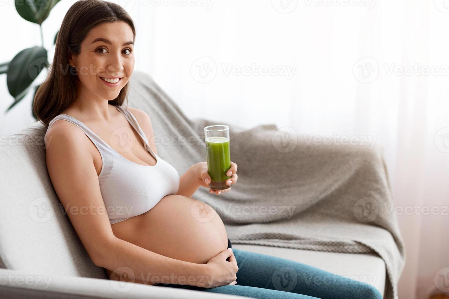 Happy Young Pregnant Woman With Glass Of Green Smoothie Sitting On Couch At Home, Smiling Expectant Woman In Bra Touching Naked Belly And Enjoying Heathy Vitamin Drink, Awaiting Lady Looking At Camera photo