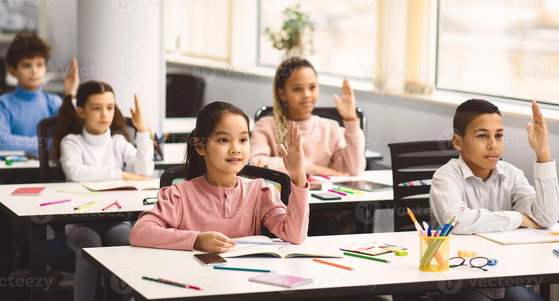 First Day Of School. Multiethnic group of diverse little classmates sitting at tables in modern classroom, raising hands up for an answer, discussing new theme with teacher, participation concept photo